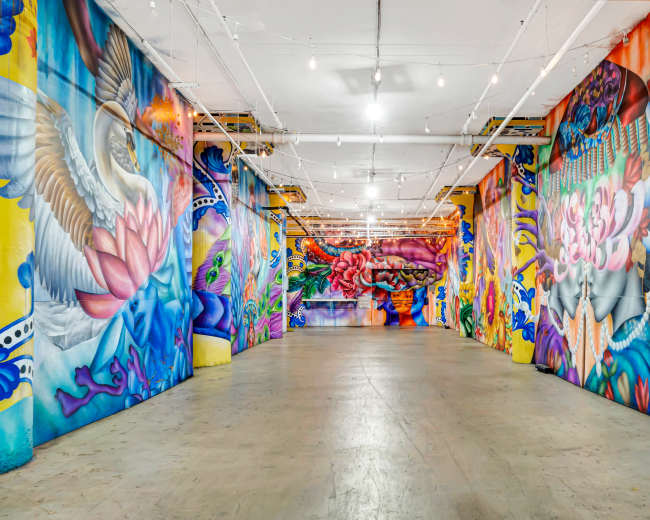 A large indoor space features walls covered in vibrant, intricate murals depicting various colorful designs and patterns.