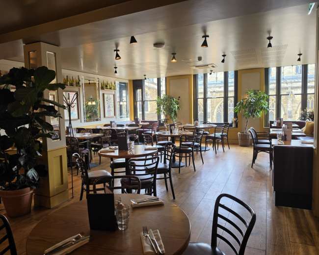 The image shows a spacious restaurant with wooden floors, several tables and chairs, and large windows letting in natural light, creating a bright atmosphere.