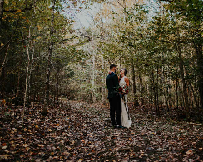 A couple stands together on a leaf-covered path in a forest, surrounded by trees.