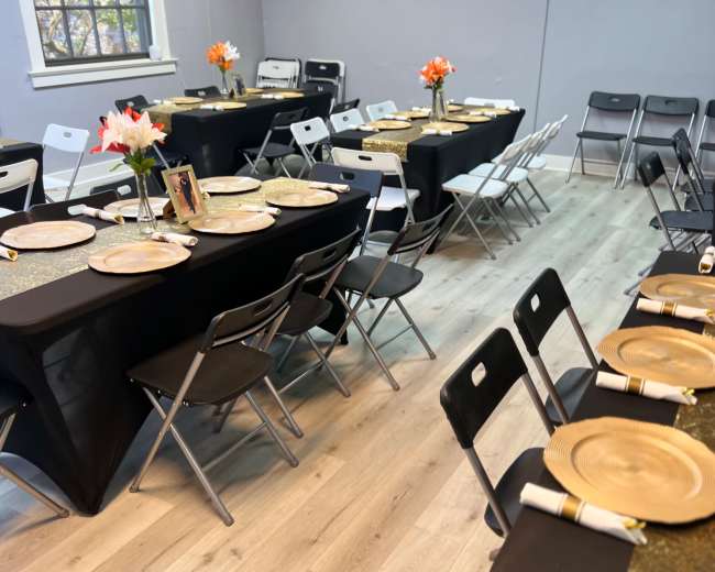 The image shows a decorated event space with several tables set for dining, featuring black tablecloths, gold plates, and flower centerpieces.