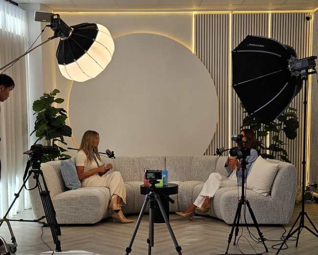 A modern interview setup features two people seated on a light-colored sofa, equipped with professional lighting and cameras in a brightly lit room.