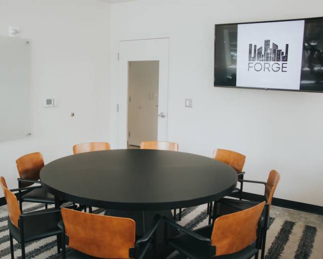 A conference room features a round black table surrounded by six wooden chairs and a wall-mounted television displaying the logo for "FORGE."