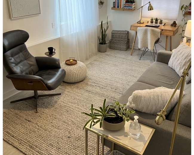 The image shows a cozy living room with a gray sofa, a black armchair, a small coffee table, and a wooden desk, all arranged around a soft rug under natural light from a window.