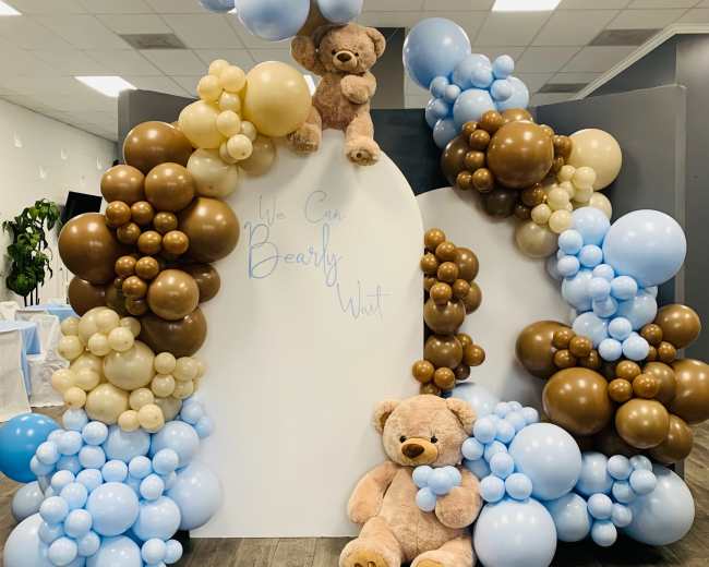 A decorative display featuring a large arrangement of balloons in various colors, including brown, cream, and light blue, along with two teddy bears positioned prominently at the center.