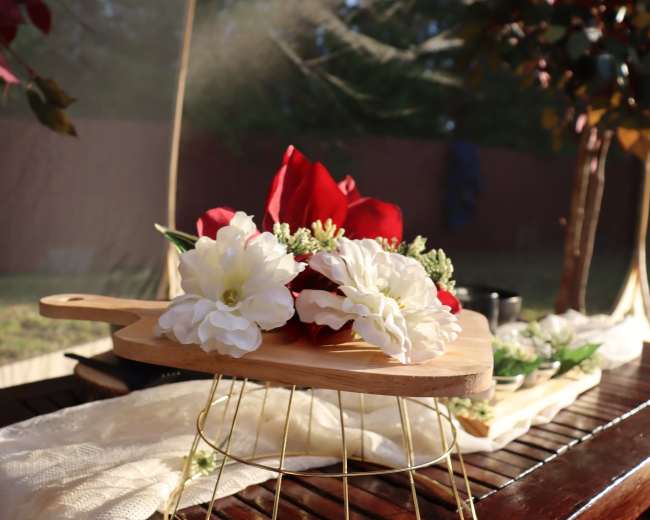 A wooden serving board adorned with white and red flowers sits on a table with a cream-colored cloth, surrounded by greenery.