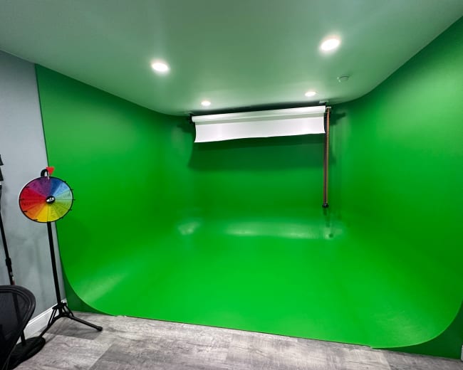 A green screen setup in a room, featuring a smooth green backdrop, overhead lighting, and a color wheel on a stand.