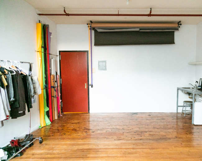 The image shows a spacious, minimally furnished studio with wooden floors, a red door, and a clothing rack filled with various garments.