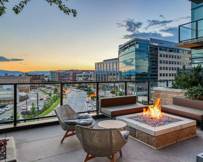 A modern balcony overlooks a city street with a fire pit surrounded by seating, set against a backdrop of buildings and a sunset sky.