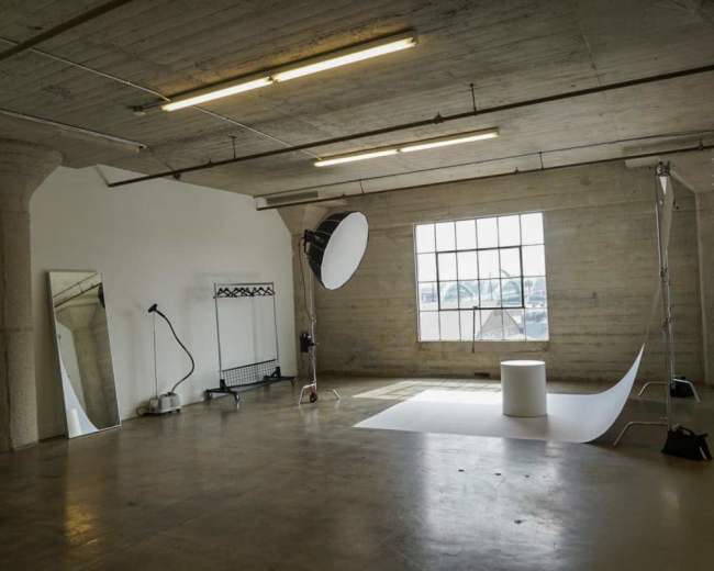 The image shows a spacious, empty studio with a large window, a backdrop setup, a mirror, and minimalist equipment.