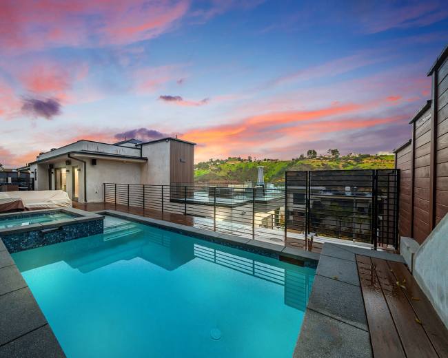 A modern rooftop pool surrounded by wooden decking and a view of hills under a colorful sunset sky.