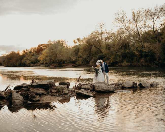 A couple in wedding attire stands on large rocks in a river, surrounded by trees as the sun sets in the background.