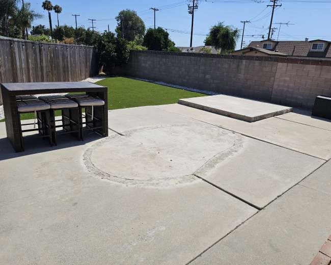 A backyard with a concrete patio, a set of four bar stools, a circular area that appears to be incomplete, and a raised platform at the far end, surrounded by a low wall and green grass.