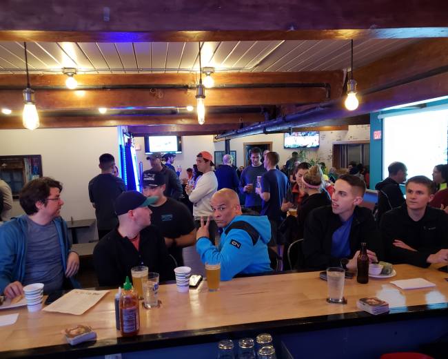 A group of people are seated at a bar with a lively atmosphere, while others socialize in the background.