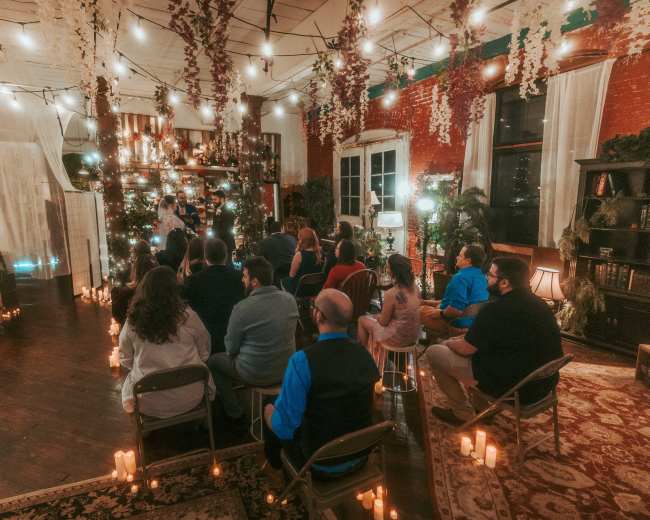 A group of people sits in chairs facing a decorated altar illuminated by string lights and candles in a vintage indoor space.
