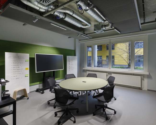 The image shows a modern conference room equipped with a round table, black chairs, a large screen, and windows overlooking a green area.