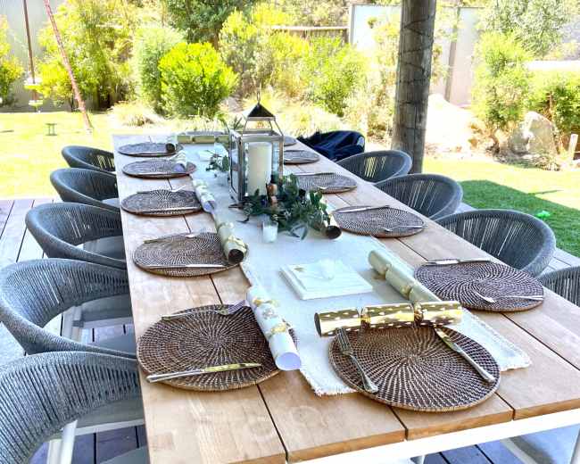 A long wooden dining table is set outdoors with round placemats and decorative items under a shaded area surrounded by greenery.