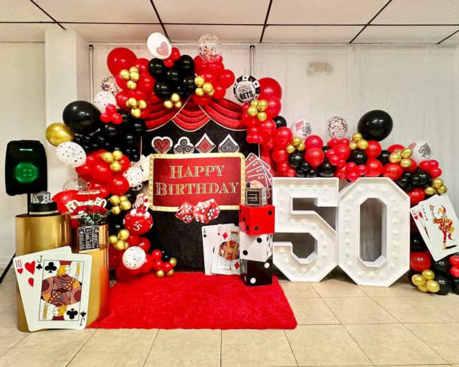 The image features a vibrant birthday celebration setup with a large sign that reads "HAPPY BIRTHDAY" and "50," surrounded by an arrangement of black, red, and gold balloons, along with playing cards and dice decorations.