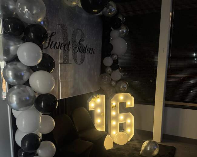 The image shows a decorated corner for a Sweet Sixteen celebration, featuring a backdrop with the text "Sweet Sixteen," large illuminated numbers "16," and an arch made of black, white, and silver balloons.