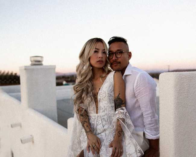 A couple poses together on a rooftop terrace, with the woman wearing a lace dress and the man in a white shirt.