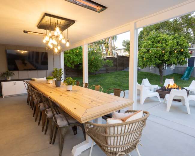 A spacious outdoor dining area features a long wooden table, surrounded by chairs, with a grassy yard and a fire pit in the background.