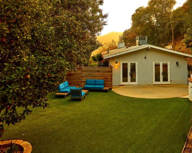 A small house with blue patio furniture sits on a green lawn surrounded by trees.