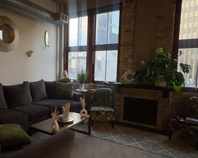 The image shows a cozy living room with large windows, a brown sectional sofa, and a fireplace beside potted plants and decorative items on a coffee table.
