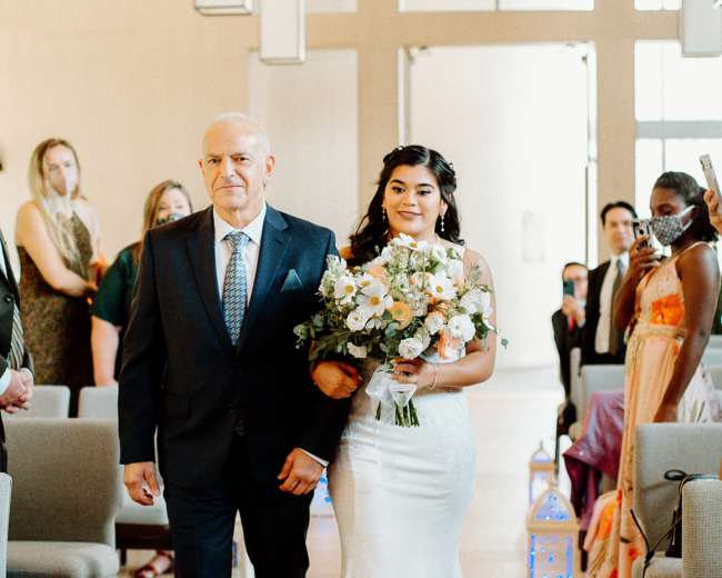 A bride in a white gown walks down an aisle accompanied by her father, as guests look on in a well-lit venue.