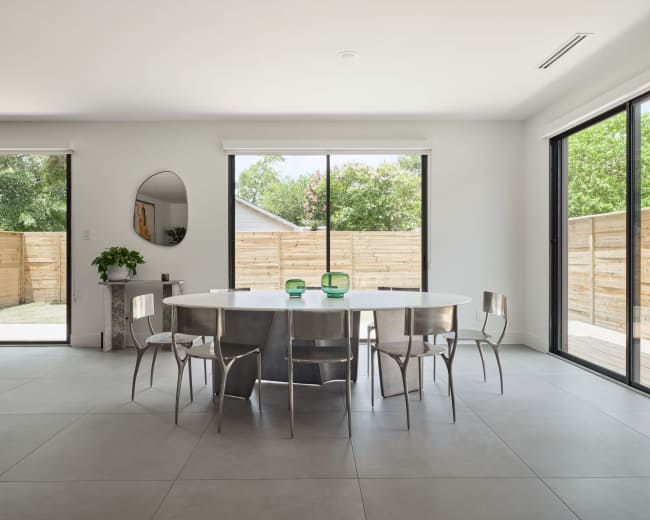 A modern dining area features a round metal table surrounded by six metal chairs, with large windows overlooking a fenced outdoor space.