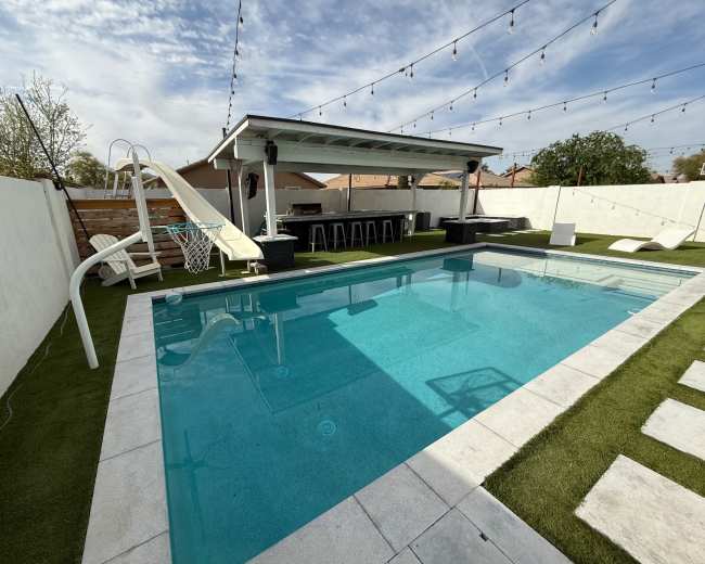 The image shows a backyard pool with a slide, a seating area under a covered bar, and decorative string lights overhead.