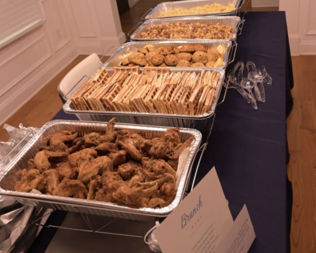 A table is set up with several trays of food for a brunch, featuring items such as fried chicken, pastries, and a variety of baked goods, accompanied by a decorative sign.