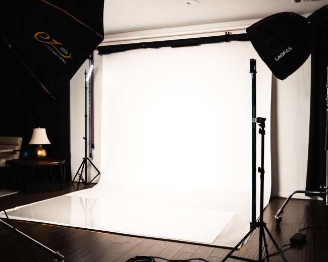 The image shows a photography studio setup with a white backdrop, professional lighting equipment, and minimal furniture.