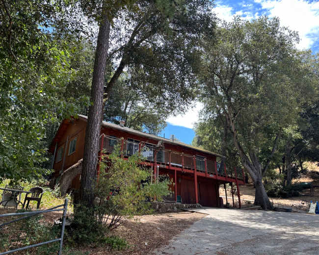 A two-story red wooden house with a balcony overlooks a gravel driveway surrounded by trees and hilly terrain.