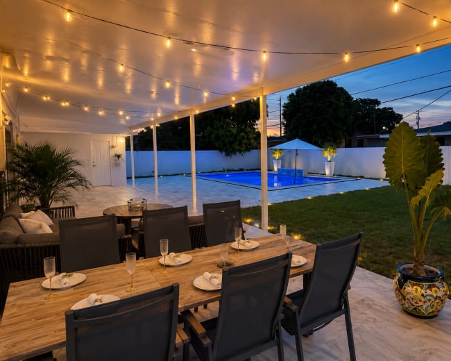 The image shows a covered outdoor dining area with a wooden table and chairs, overlooking a well-lit pool and a grassy yard at dusk.