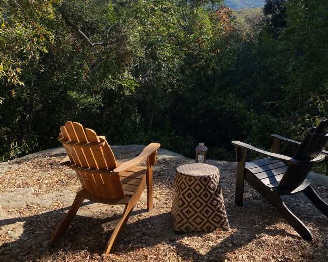 Two chairs sit on a stone patio surrounded by lush greenery, facing a scenic view of distant mountains under a clear blue sky.
