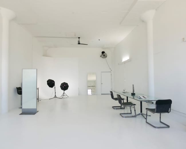 The image shows a bright, empty studio space featuring a long glass table, several black chairs, and photography equipment.