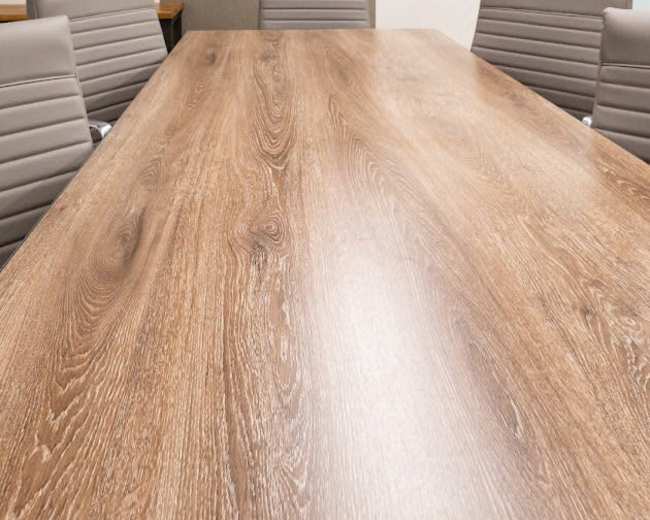 A long wooden conference table is surrounded by several gray chairs in a modern meeting room.