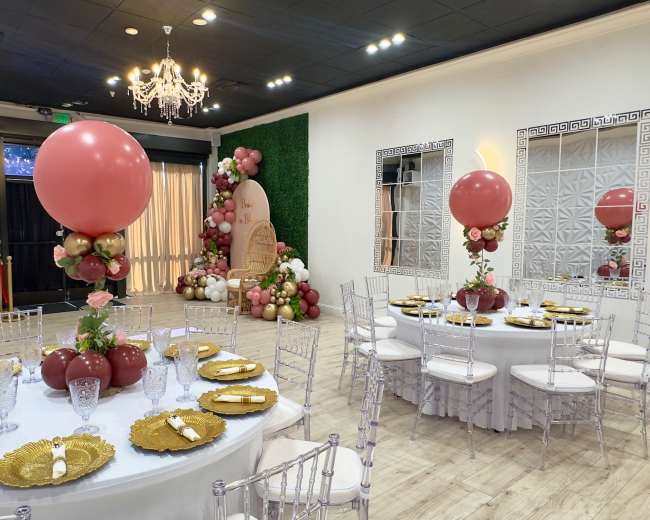 The scene shows a decorated banquet room set up for an event, featuring round tables adorned with golden tableware and large pink balloon centerpieces.