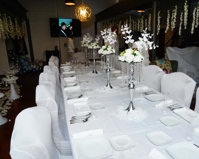 A long, elegantly set dining table with white decorations, featuring floral centerpieces and a television playing in the background.