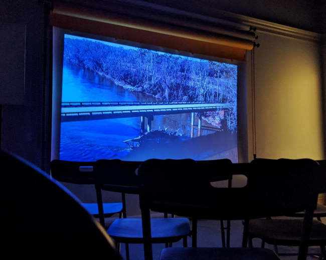 A projection of a river and bridge scene is displayed on a screen in a dimly lit room with empty chairs arranged in front of it.