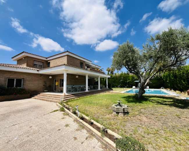 The image shows a large two-story house with a patio area, surrounded by a grassy yard and a swimming pool, under a partly cloudy sky.