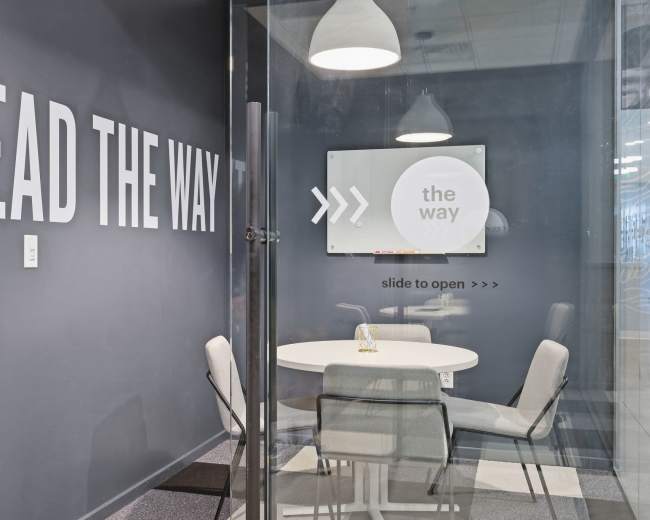 The image shows a modern meeting room with a round table and four chairs, featuring glass walls and a wall with large text that reads "LEAD THE WAY."