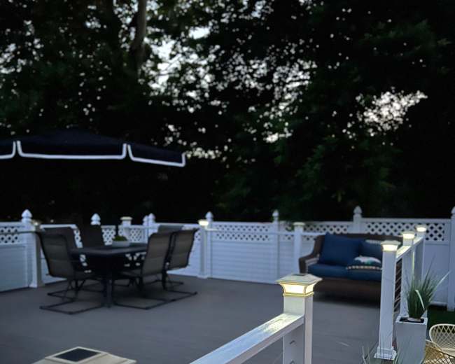 The image shows a well-lit deck with a dining set and a lounge area, surrounded by greenery at dusk.