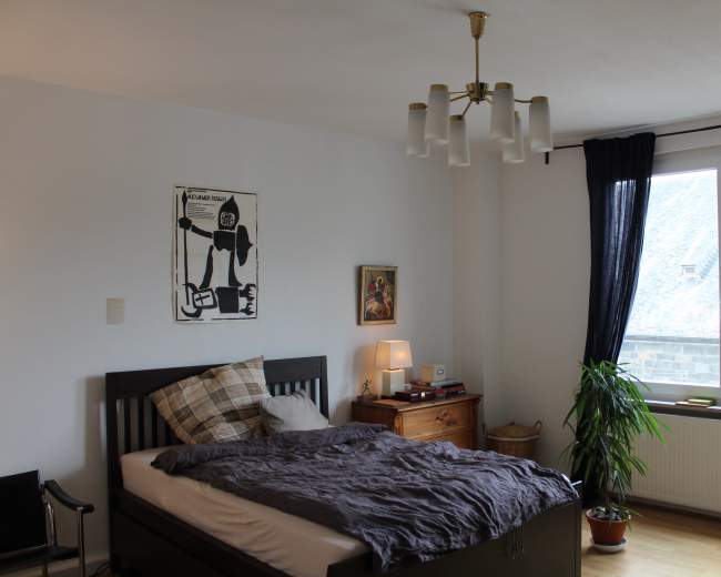 The bedroom features a bed with a dark gray duvet, a bedside lamp, a wooden nightstand, a chair, and a large window with dark curtains, illuminated by natural light.