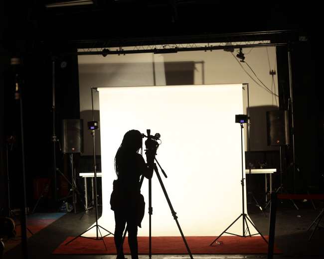 A silhouette of a person stands with a camera in front of a lit white backdrop in a photography studio.