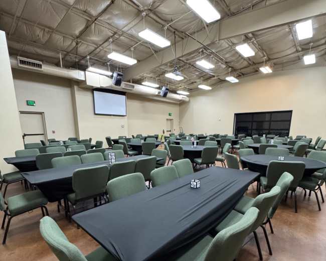 The image shows a large, empty banquet hall with multiple rows of tables covered in black tablecloths and green chairs arranged around them.