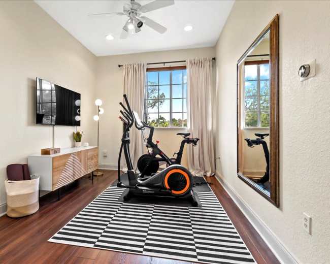 A home gym featuring an elliptical machine, a large mirror, a TV mounted on the wall, and a striped rug on a wooden floor.