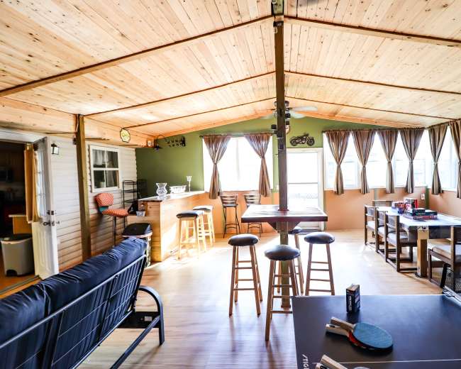 The image depicts a spacious indoor sunroom with a wooden ceiling, a bar area, seating, and large windows allowing natural light.
