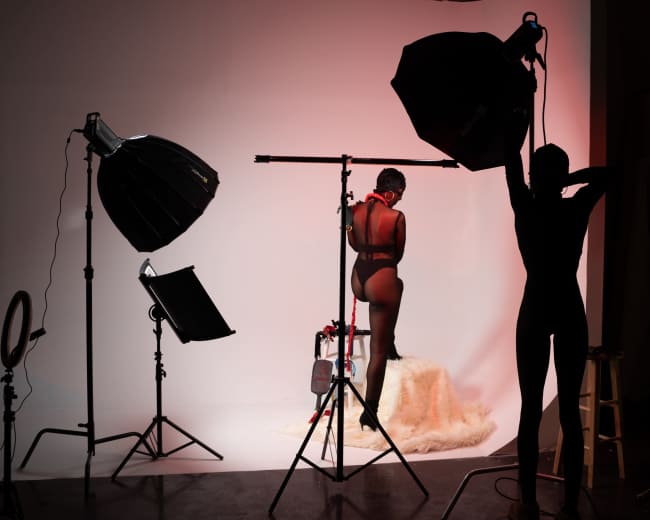 A woman poses on a stool in a photography studio, surrounded by lighting equipment against a plain backdrop.