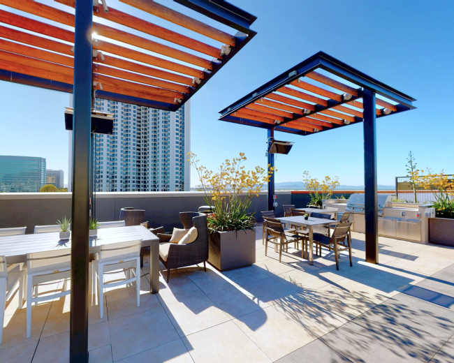 The image shows a rooftop terrace featuring seating areas, shaded pergolas, and planters with plants, alongside a city skyline in the background.
