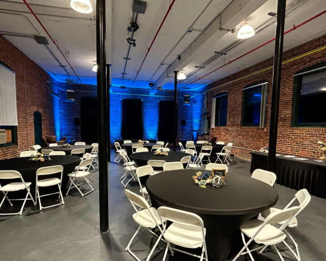 A spacious event venue with several round tables, each covered in black tablecloths, arranged in a well-lit area featuring blue lighting on the brick walls.
